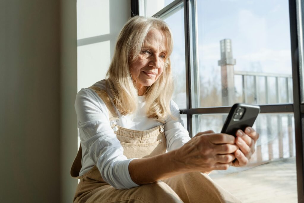 pexels photo 7330716 7330716 1 Elderly woman using a smartphone by a window, enjoying leisure time indoors.